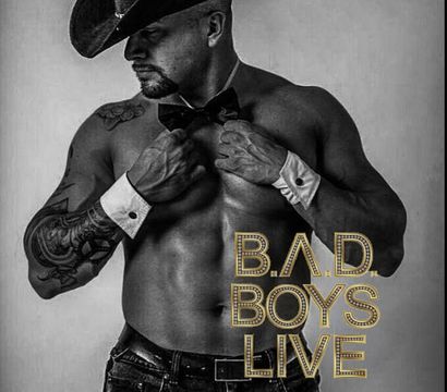 Black-and-white promo poster of a shirtless, muscular male performer wearing a cowboy hat, bow tie and wrist cuffs, showing tattoos and jeans, posing alongside gold marquee text 'B.A.D. BOYS LIVE'.