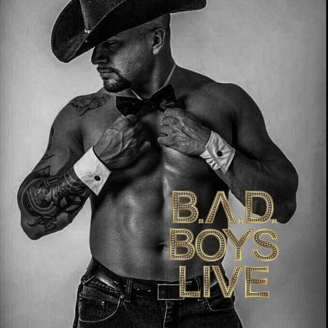 Black-and-white promo poster of a shirtless, muscular male performer wearing a cowboy hat, bow tie and wrist cuffs, showing tattoos and jeans, posing alongside gold marquee text 'B.A.D. BOYS LIVE'.