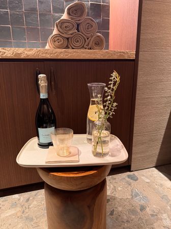 Spa lounge scene with a tray on a wooden stool holding a bottle of sparkling wine, a glass, a lemon-slice carafe and a small vase with greenery, stacked rolled towels on a stone counter and warm wood cabinetry in the background.