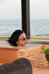 Person in sunglasses relaxing in an outdoor wooden hot tub at a beachfront with wicker accents and calm ocean horizon in the background.