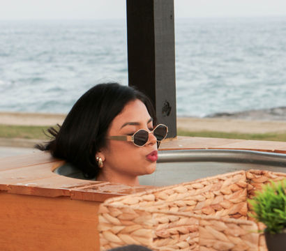 Person in sunglasses relaxing in an outdoor wooden hot tub at a beachfront with wicker accents and calm ocean horizon in the background.