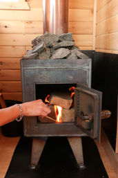 Hand lighting a wood-burning stove in a wood-paneled cabin, flames glowing inside and sauna stones stacked on the metal heater.
