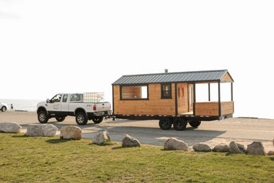White pickup truck towing a wooden tiny-house trailer parked at a rocky seaside parking area with the ocean in the background.