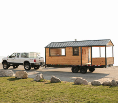 White pickup truck towing a wooden tiny-house trailer parked at a rocky seaside parking area with the ocean in the background.