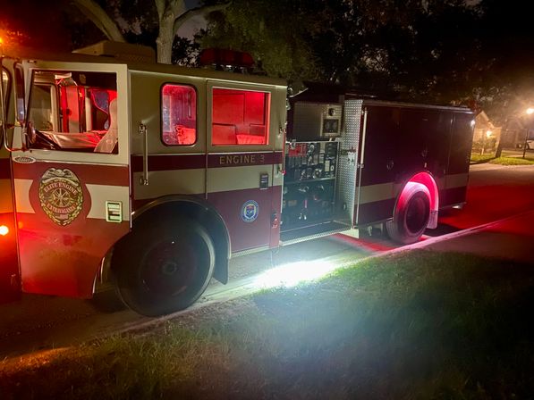 Red fire truck parked on a suburban street at night, red emergency lights glowing and an open equipment panel casting light onto the curb and grass.