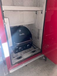 Compact portable gas grill stored inside a red vehicle side compartment on a diamond-plate step, ready for outdoor grilling