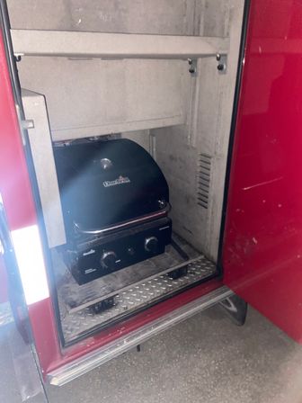 Compact portable gas grill stored inside a red vehicle side compartment on a diamond-plate step, ready for outdoor grilling