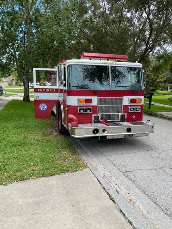 Red-and-white fire truck labeled Engine 3 parked curbside on a tree-lined suburban street, front view with open door, sidewalk and green lawns