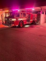 Red fire engine parked under a fire station canopy at night, purple-pink emergency lights casting colorful reflections on wet asphalt.