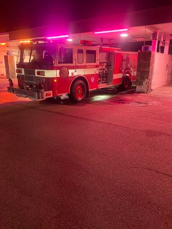Red fire engine parked under a fire station canopy at night, purple-pink emergency lights casting colorful reflections on wet asphalt.