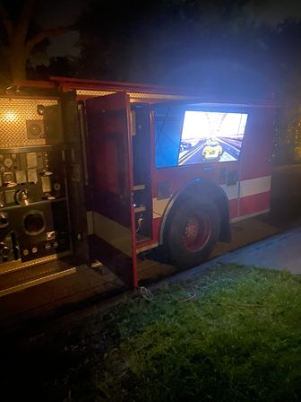 Red fire truck parked on a residential street at night with an open side compartment revealing a flat-screen TV glowing blue and showing a racing video game.