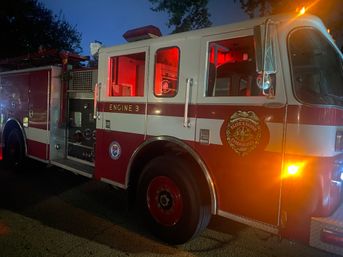 Red and white fire engine parked on a dim street at dusk, side view showing Engine 3 lettering, open equipment compartments, glowing red interior and amber emergency lights.