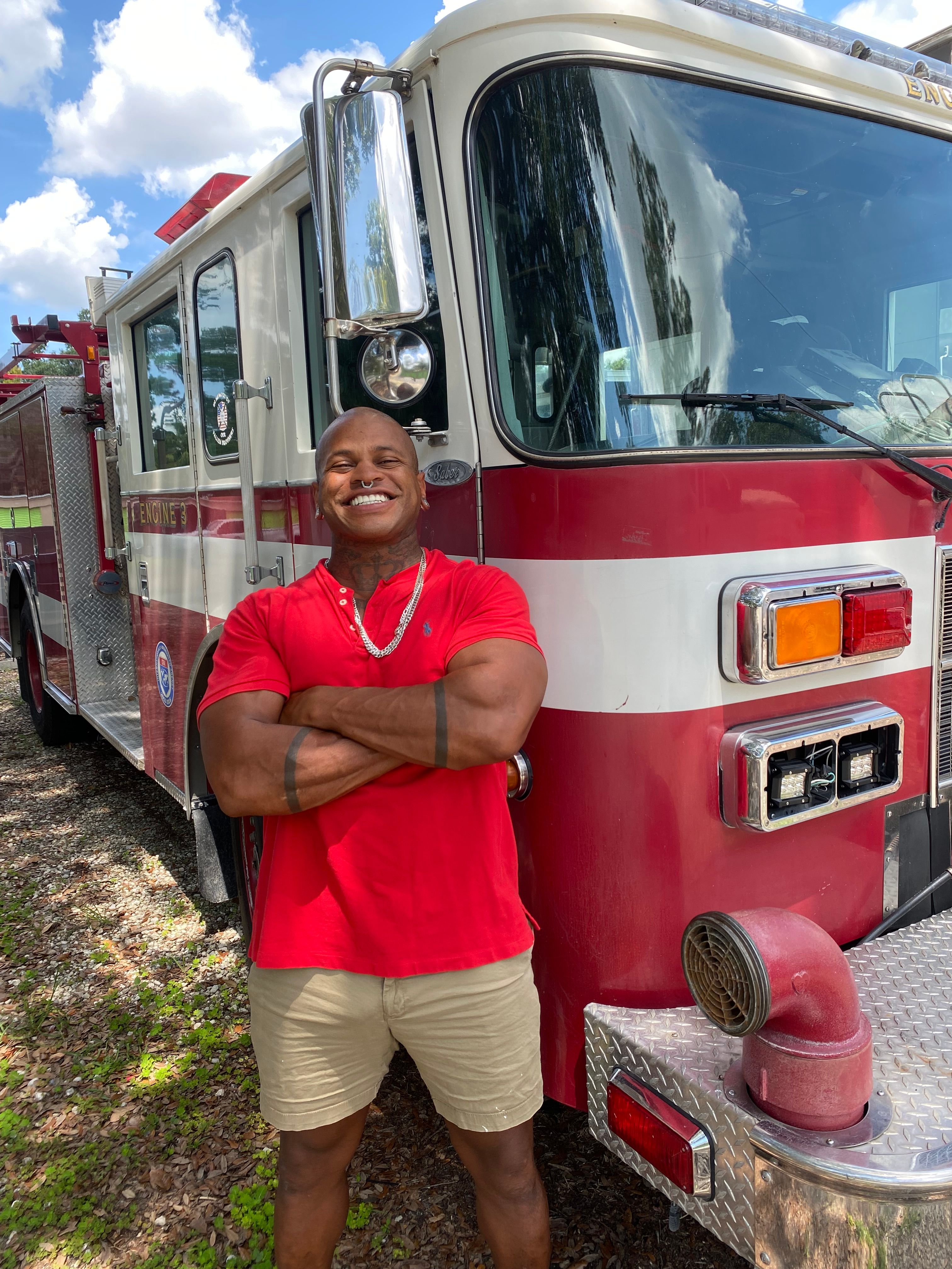 Smiling man in a red polo and khaki shorts stands with arms crossed beside a red fire engine under a bright blue sky.
