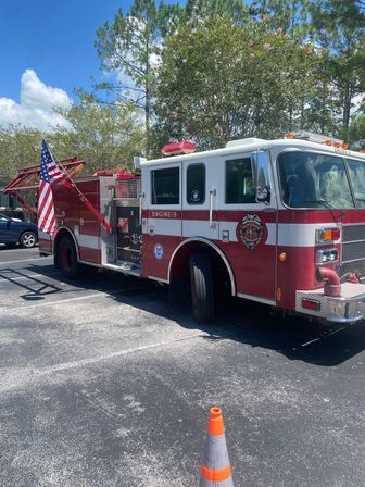 Patriotic red-and-white fire engine (Engine 3) parked in a sunny suburban parking lot with an American flag, orange safety cone, and tall pine and flowering trees under a bright blue sky