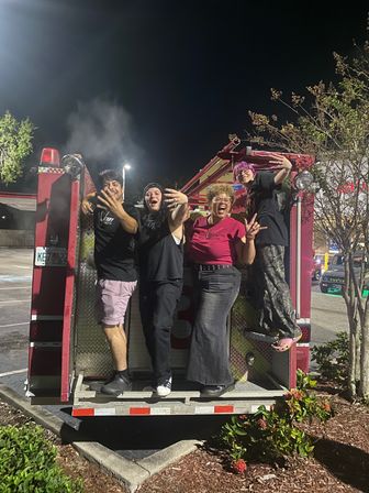 Four friends posing on the back of a red fire truck at night in a parking lot, laughing and making playful hand gestures under streetlights.