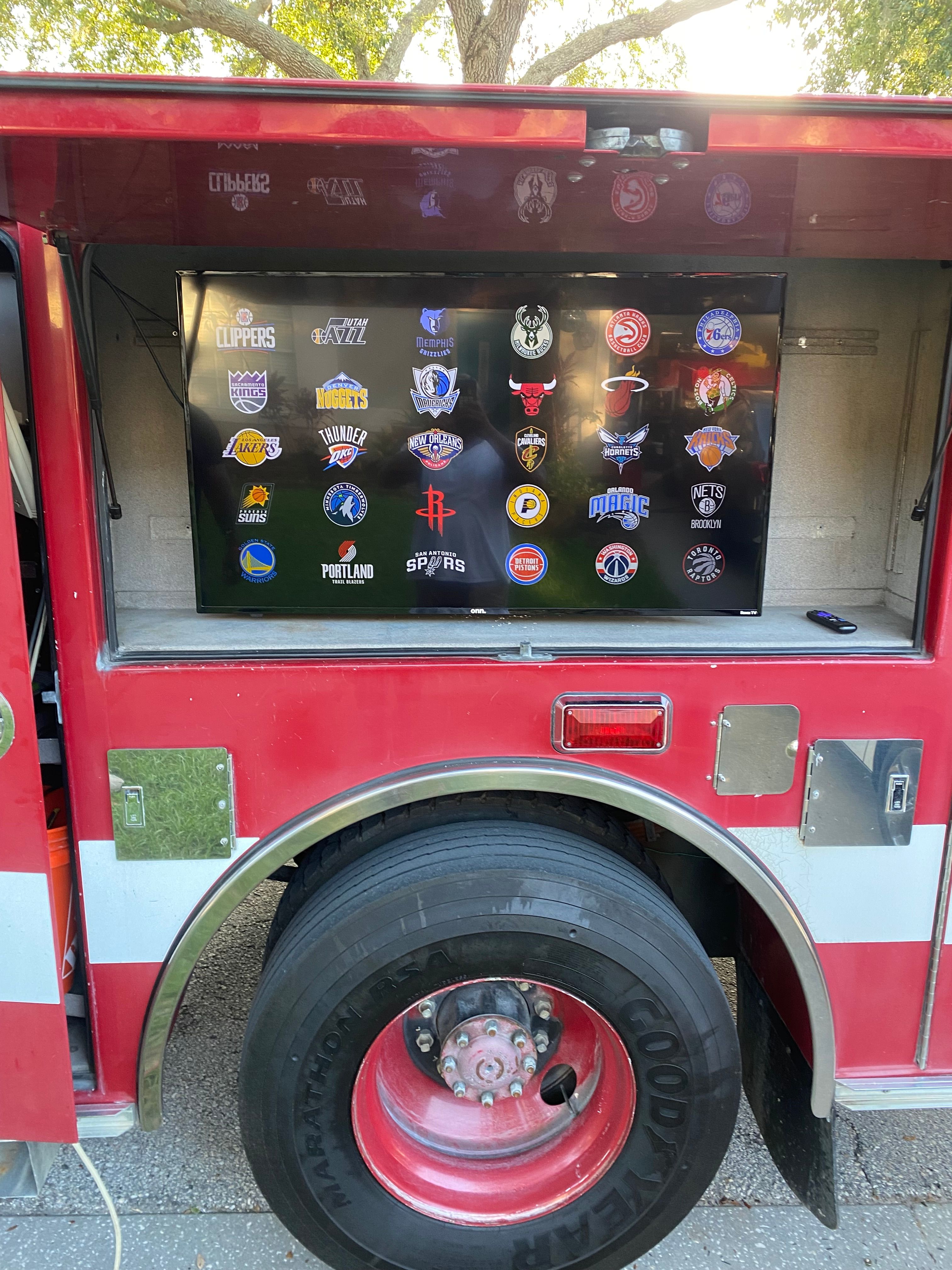 Red fire truck side compartment opened to reveal a flat-screen TV showing NBA team logos, mounted above a large red truck wheel, parked outdoors under a tree.