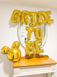 Gold foil balloons spelling BRIDE TO BE and a Mickey-ear gold balloon reflected in an oval mirror above a wooden console table — bridal shower/bachelorette décor in a bright entryway