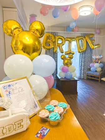 Bridal shower setup in a living room with gold letter balloons spelling BRIDE, gold ear-shaped foil balloons, pastel pink, blue and white balloon clusters, a table with a framed welcome sign and pink and turquoise frosted cupcakes.