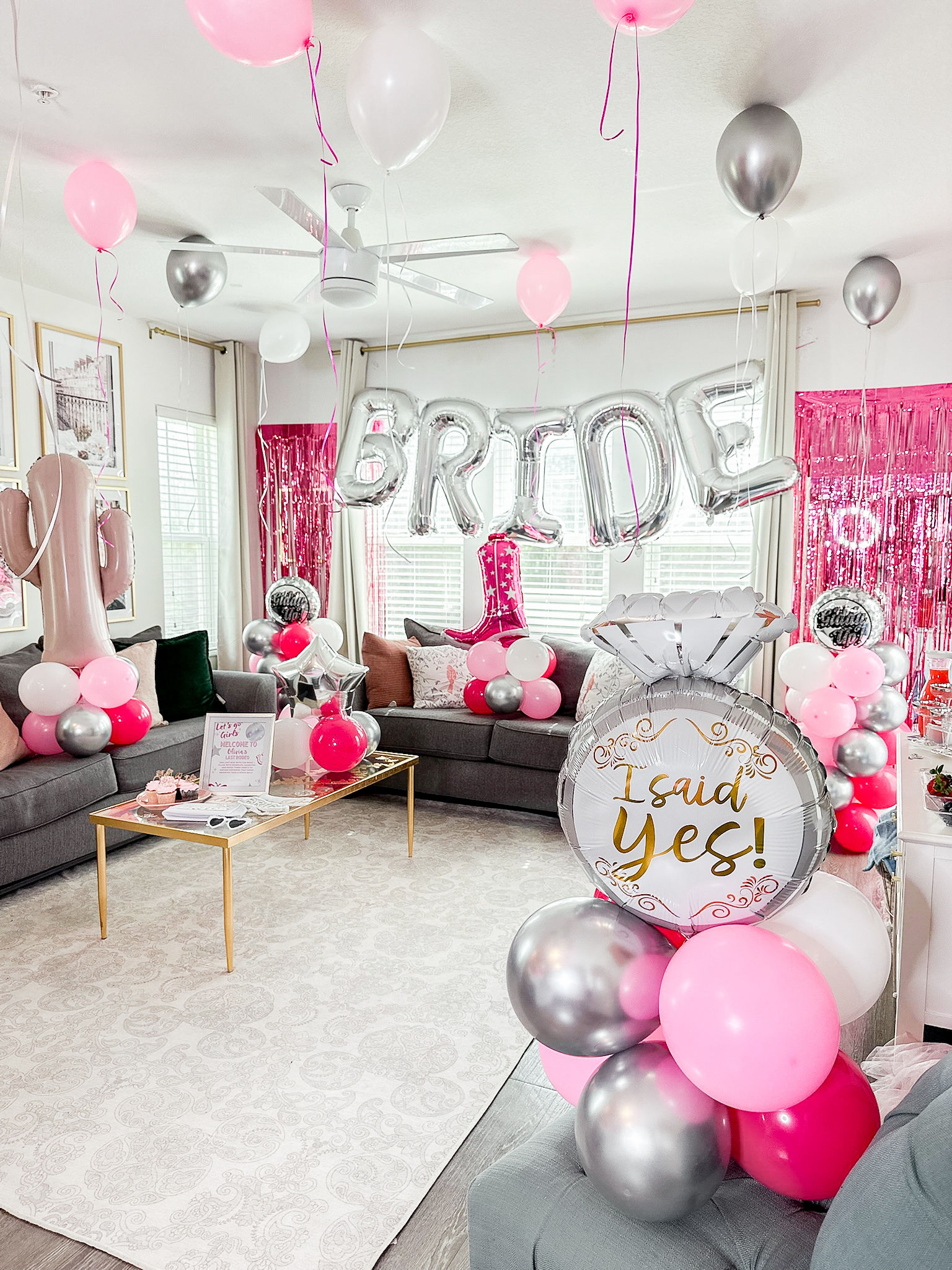 Bright home living room bridal shower decorations with silver 'BRIDE' letter balloons, pink, white and metallic silver balloon clusters, a large 'I said yes!' ring balloon, pink fringe backdrop and gray sofas.