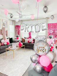 Bright home living room bridal shower decorations with silver 'BRIDE' letter balloons, pink, white and metallic silver balloon clusters, a large 'I said yes!' ring balloon, pink fringe backdrop and gray sofas.