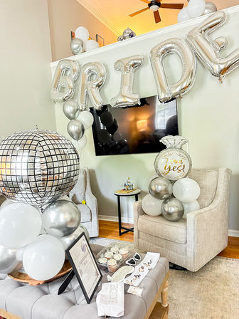 Silver-and-white living room bridal shower setup with foil letter 'BRIDE' balloons, a 'I said yes' ring balloon, disco-ball balloon cluster, cupcake tray and 'bride-to-be' sash on an ottoman.