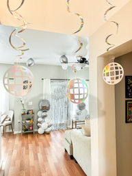 Bright living room party setup with holographic hanging disco ball cutouts, curled metallic streamers, silver-and-white balloon columns and a metallic fringe photo backdrop over hardwood floors.