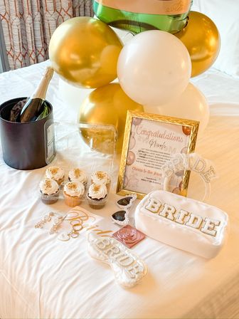 Bachelorette setup on a hotel bed with gold-and-white balloons, champagne in an ice bucket, cupcakes, framed congratulations sign and 'BRIDE' accessories