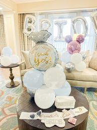 Bright hotel suite styled for a bridal celebration with pastel blue, white and silver balloon clusters, a large foil ring balloon reading 'I said yes!', silver 'BRIDE' letter balloons, and bride-to-be accessories (sash, eye mask, pouch) on a round wooden table.