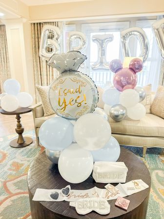 Bright hotel suite styled for a bridal celebration with pastel blue, white and silver balloon clusters, a large foil ring balloon reading 'I said yes!', silver 'BRIDE' letter balloons, and bride-to-be accessories (sash, eye mask, pouch) on a round wooden table.