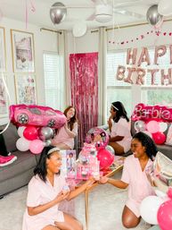 Barbie-themed birthday in a bright living room: four women in matching pink pajamas surrounded by pink and silver balloons, Barbie-shaped balloons, foil "Happy Birthday" letters, a pink fringe backdrop and cupcakes.
