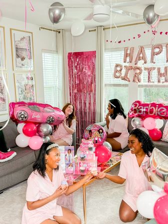 Barbie-themed birthday in a bright living room: four women in matching pink pajamas surrounded by pink and silver balloons, Barbie-shaped balloons, foil "Happy Birthday" letters, a pink fringe backdrop and cupcakes.