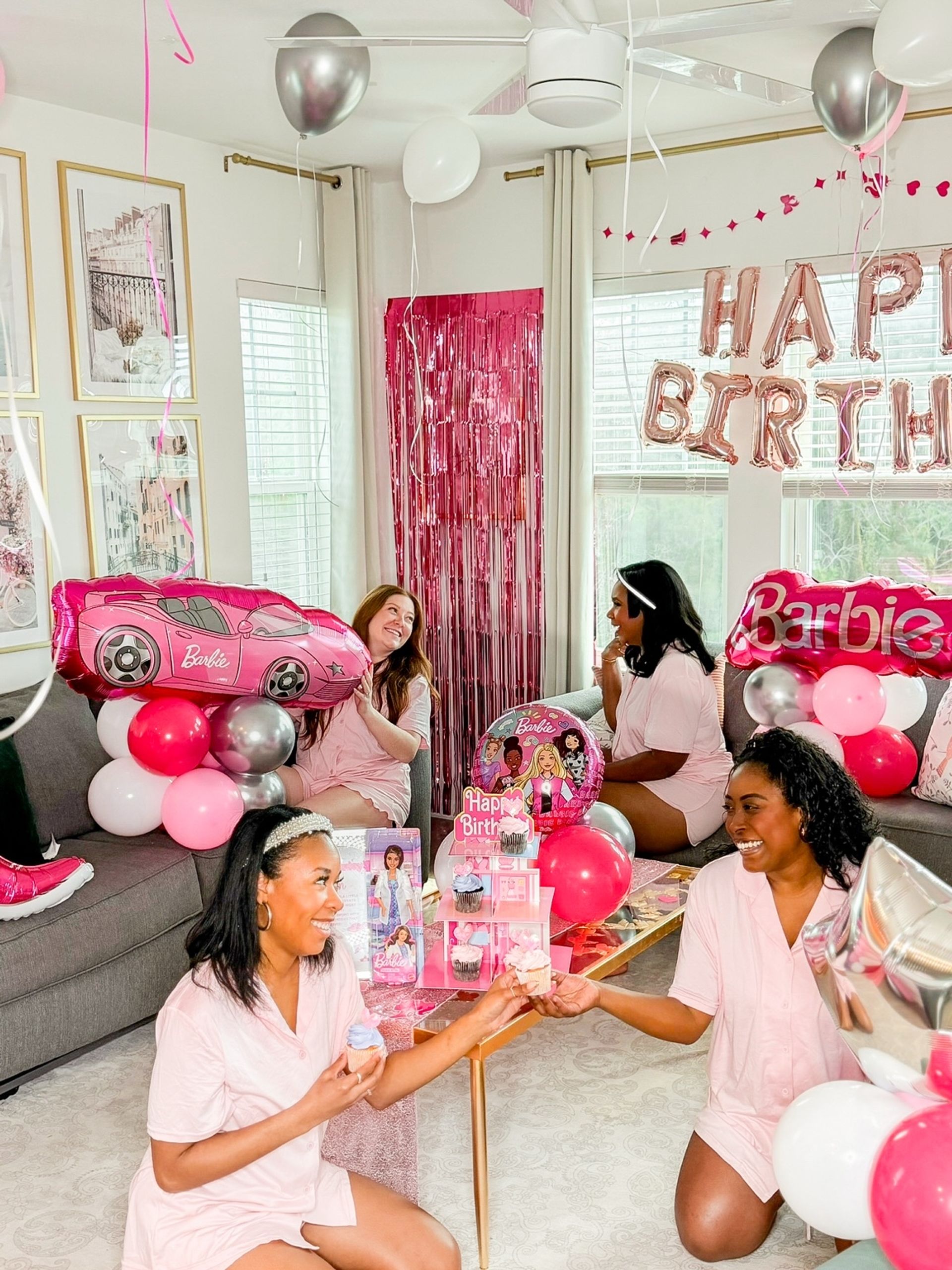 Barbie-themed birthday in a bright living room: four women in matching pink pajamas surrounded by pink and silver balloons, Barbie-shaped balloons, foil "Happy Birthday" letters, a pink fringe backdrop and cupcakes.