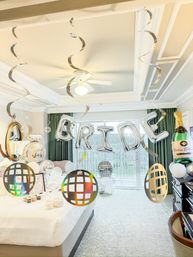 Bright hotel bridal suite decorated in white and silver for a bridal celebration with large silver 'BRIDE' balloons, champagne bottle balloon, metallic hanging spirals and disco-ball ornaments above the bed and balcony.