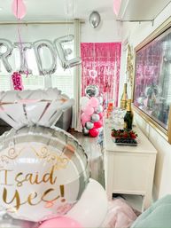 Cozy living room engagement party with pink and silver balloons, an 'I said yes!' diamond balloon, silver letter balloons spelling BRIDE, pink tinsel backdrop, champagne bottles and a fruit platter on a white sideboard.