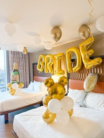 Hotel bridal suite decorated with gold letter balloons spelling BRIDE, white and gold balloon clusters and champagne foil balloons above two beds.