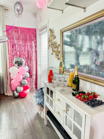 Bright pink-themed indoor party scene with a metallic fringe backdrop and balloon column next to a white sideboard styled as a mimosa bar with champagne bottles, juice carafes, stacked coupe glasses and trays of strawberries, blueberries and blackberries.