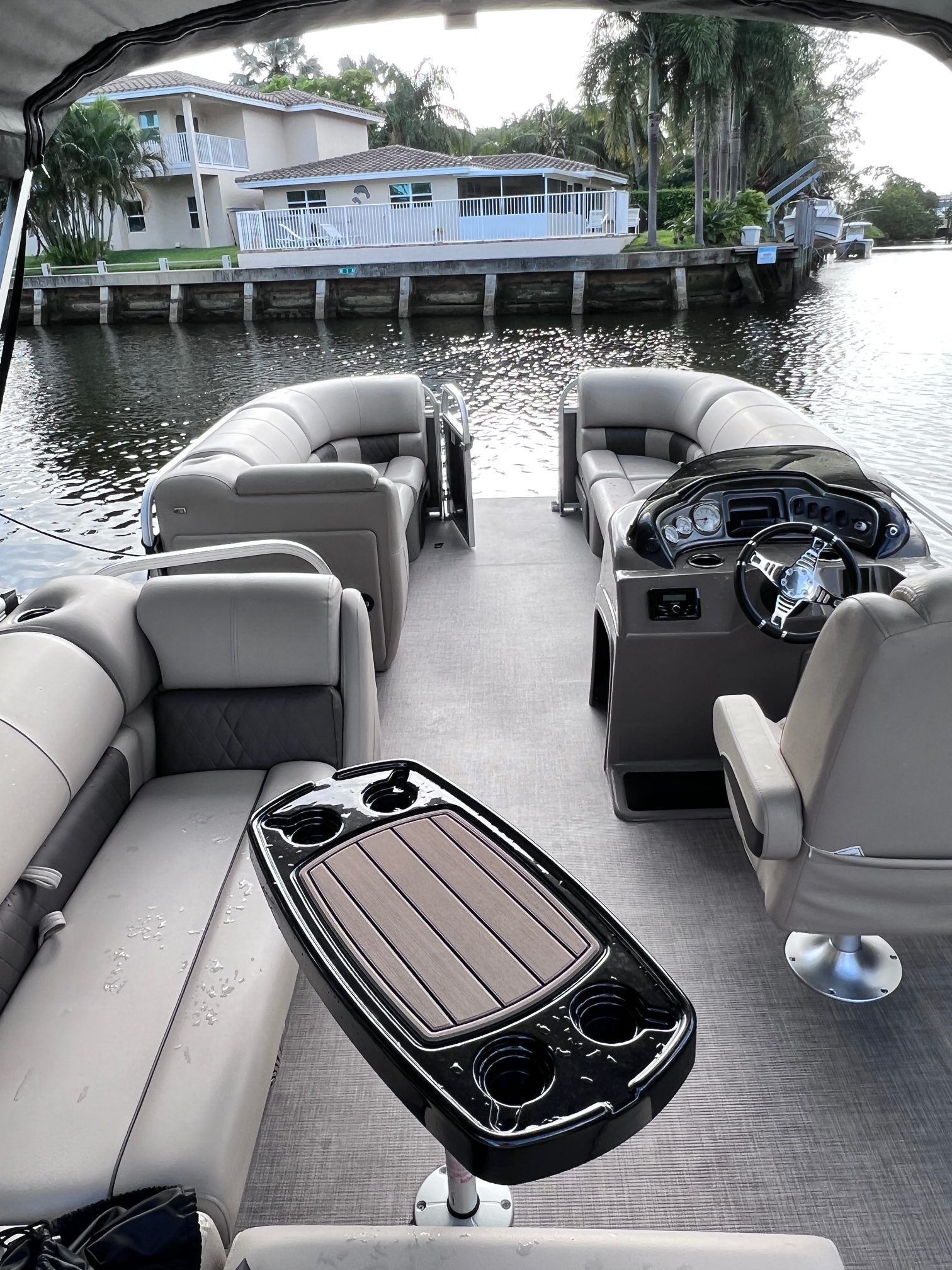 Beige pontoon boat interior with helm, curved seating and a table with cupholders, docked along a residential waterfront canal with palm trees and houses