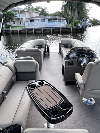 Beige pontoon boat interior with helm, curved seating and a table with cupholders, docked along a residential waterfront canal with palm trees and houses
