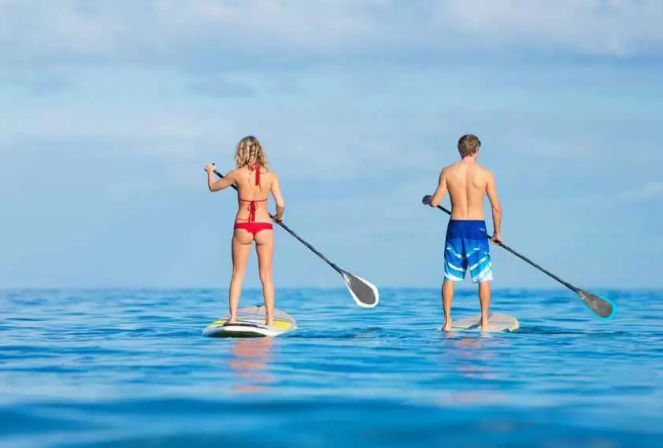 Two paddleboarders standing on SUP boards, paddling across calm turquoise ocean under a clear blue sky — sunny summer water activity