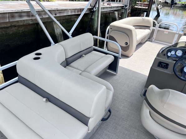 Beige pontoon boat interior with cushioned bench seating, dual cup holders, helm console and textured flooring, docked alongside a wooden waterfront pier