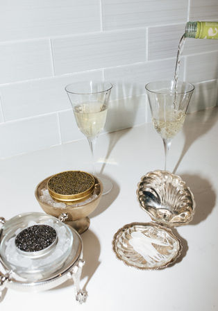 Sparkling white wine poured into flutes on a white subway-tiled kitchen counter beside tins of black and golden caviar on ice with mother-of-pearl spoons in a shell-shaped silver dish.