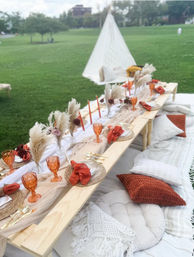 Boho outdoor picnic on a green park lawn with a low wooden table, rust-orange goblets and napkins, woven chargers, pampas grass centerpieces, gold flatware, cozy pillows and a white teepee in the background.