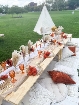 Boho outdoor picnic on a green park lawn with a low wooden table, rust-orange goblets and napkins, woven chargers, pampas grass centerpieces, gold flatware, cozy pillows and a white teepee in the background.