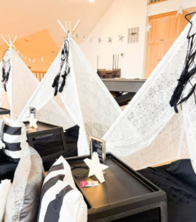 Cozy indoor kids' teepee sleepover setup in a loft with white lace teepees, hanging dreamcatchers, black-and-white striped pillows and low black tray tables topped with star decorations.