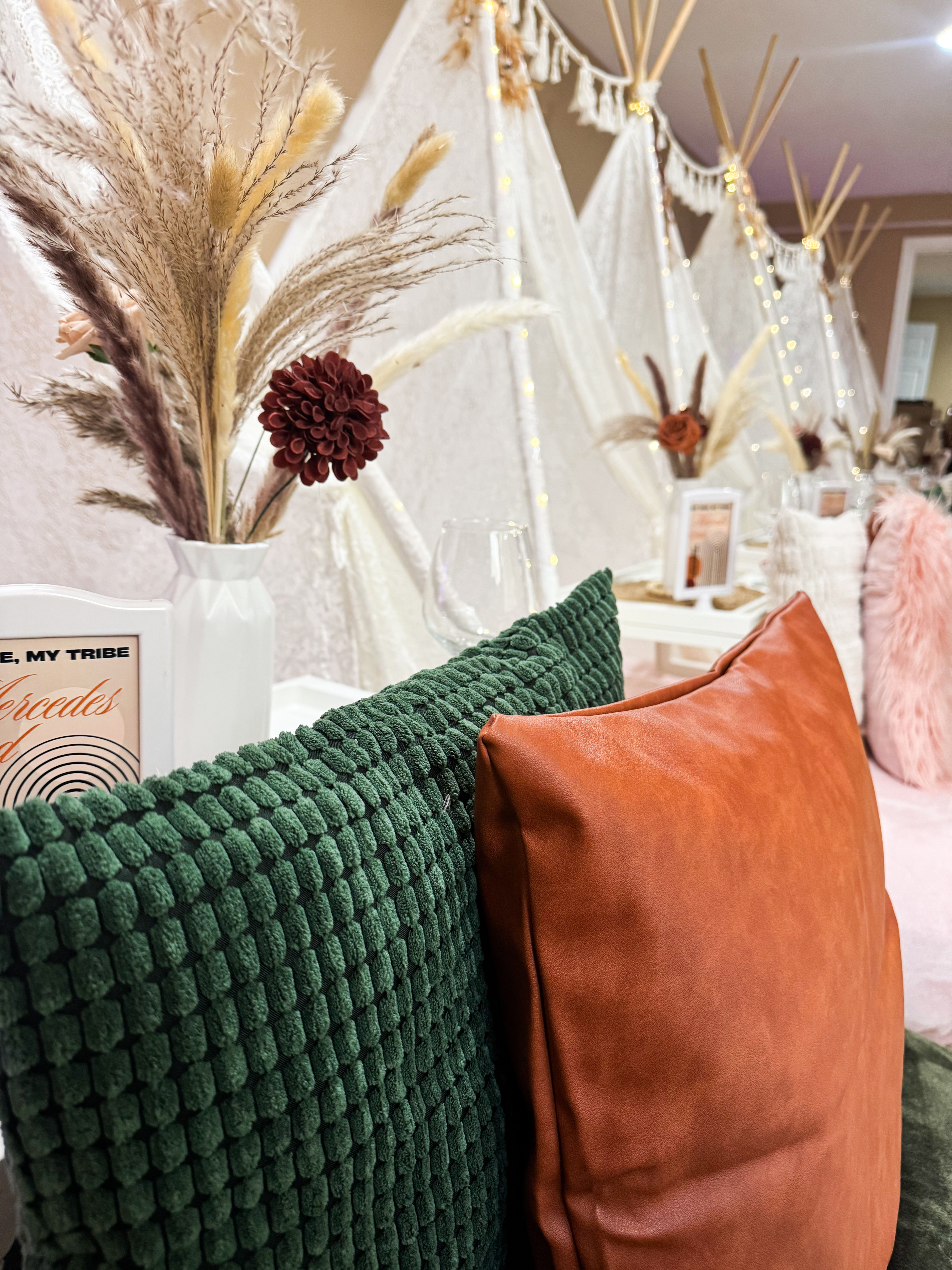 Cozy boho-chic indoor lounge with green textured and rust leather pillows, pampas grass in vases, white teepee tents and warm fairy lights
