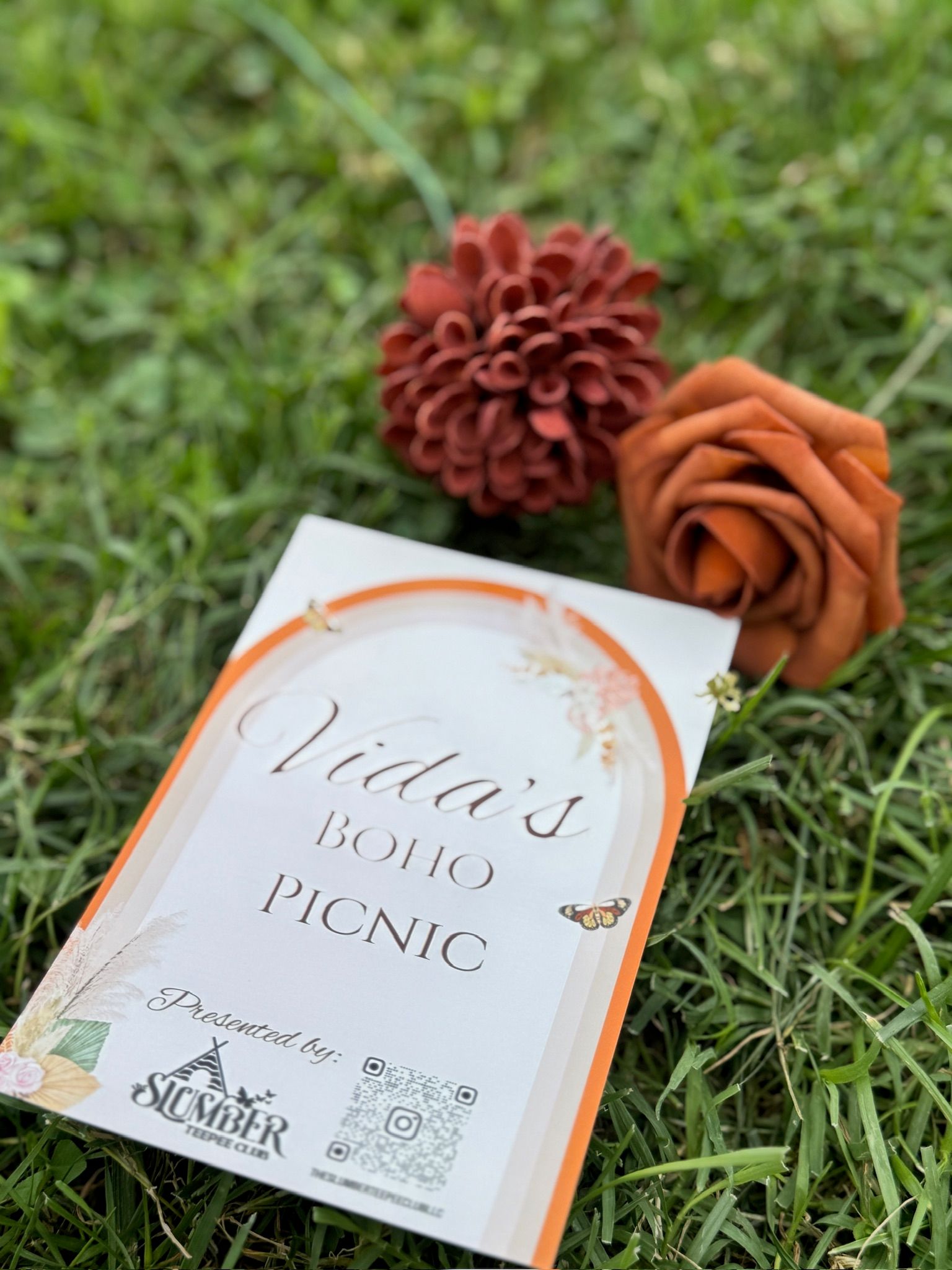 Boho picnic invitation card with orange border lying on green lawn beside two rust-colored faux flowers and a small butterfly graphic.