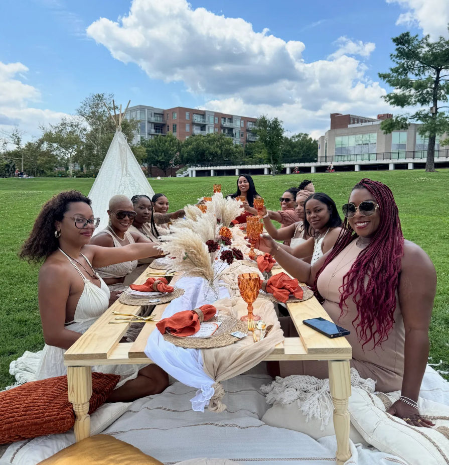 Group of women toasting at a boho-style outdoor picnic around a low wooden table with pampas grass, amber goblets and orange napkins in an urban park under a blue sky