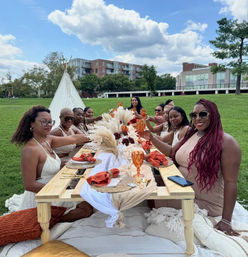 Group of women toasting at a boho-style outdoor picnic around a low wooden table with pampas grass, amber goblets and orange napkins in an urban park under a blue sky