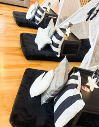 Cozy indoor kids slumber party setup: row of black plush floor beds on hardwood, striped black-and-white pillows, star-shaped and silver sequin cushions, small black side tables and white canopy teepees.