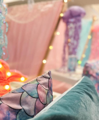 Indoor pastel play nook with a teal velvet cushion and mermaid-scale pillow in front of a pink fabric tent lit by warm string lights and soft hanging jellyfish decorations.